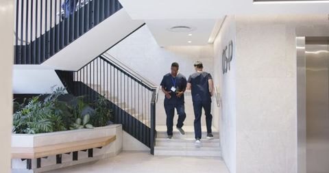 Diverse healthcare coworkers walking stairs in modern lobby checking badge and phone in navy scrubs