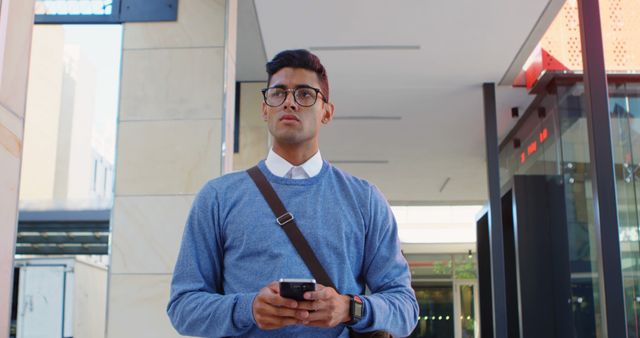 Young professional man wearing glasses and casual attire using a smartphone on a city street. Ideal for topics related to urban living, modern technology, or business communication.