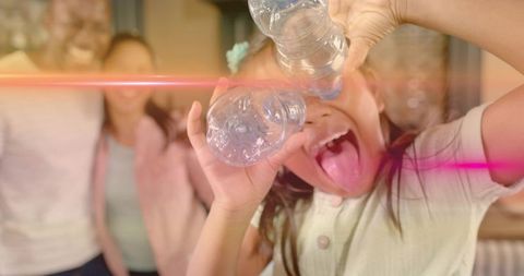Child playing with water bottles sticking out tongue while family laughing in cozy kitchen