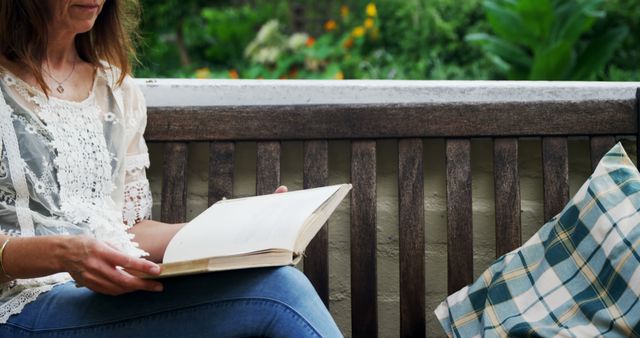Woman enjoying a quiet moment reading a book on a wooden bench in a lush garden. Suitable for themes of relaxation, leisure, peacefulness, and personal time. Ideal for use in articles or content about reading, outdoor activities, or serene environments.