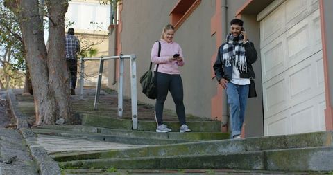 Diverse friends walking down mossy hillside stairs while checking phones, casual urban lifestyle