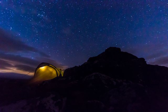 Camping tent glowing under starry sky in mountainous wilderness at night. Perfect for illustrating adventure travel, stargazing, outdoor activities, and the beauty of remote landscapes. Ideal for use in travel blogs, outdoor magazines, and nature-focused websites.