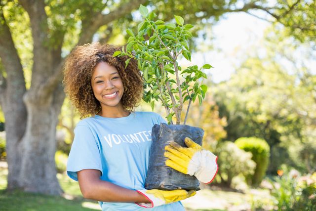 Smiling woman volunteer holding a plant in a park, wearing gloves and casual clothing. Ideal for use in community service promotions, environmental campaigns, gardening events, and volunteer recruitment materials.