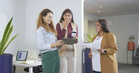 Diverse female coworkers collaborating in modern office sharing tablet and documents with coffee