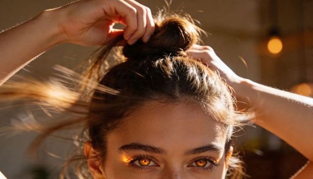 Young woman tying hair into top bun with glowing amber eyes and warm bokeh light