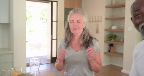 Mature couple sharing sunlit kitchen moment, woman playfully gesturing near wooden table