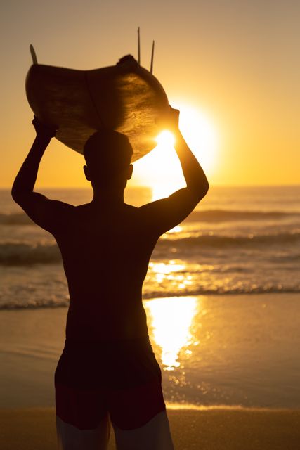 Silhouette of a man holding a surfboard on his head while standing on the beach at sunset. Ideal for use in travel brochures, surfing magazines, summer vacation promotions, and lifestyle blogs focusing on outdoor activities and adventure sports.