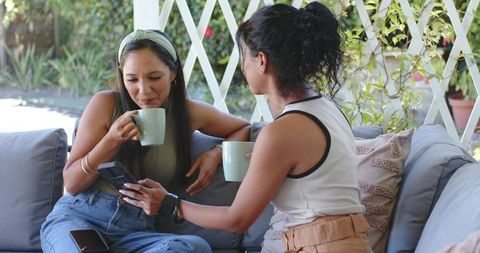 Two friends sipping coffee sharing smartphone on sunny cozy garden patio sofa