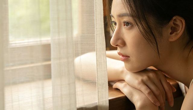 Asian woman gazing through sheer curtain leaning on wooden sill resting in soft warm light