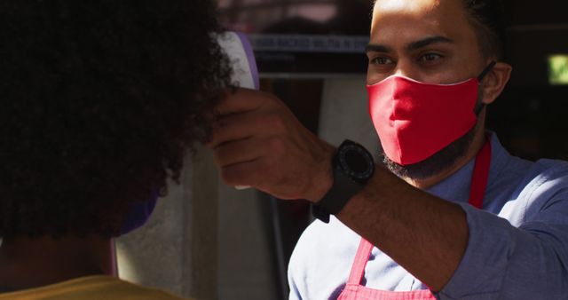 Image showing a restaurant worker wearing protective face mask and apron while checking the temperature of a customer. This scene represents health and safety measures being taken in public spaces during the COVID-19 pandemic. Ideal for use in articles and campaigns related to public health, safety protocols, customer service, and the hospitality industry.