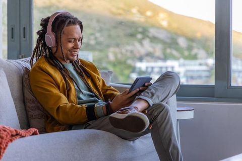 African american man relaxing on sofa with headphones using tablet by sunny hillside window