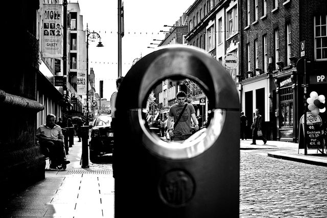 This artistic urban scene in black and white captures daily life on a cobblestone street viewed through a circular object, creating a unique perspective. It features people walking, sitting outdoors, and buildings with diverse signage. Ideal for concepts in lifestyle, city life, urban exploration, and street photography.