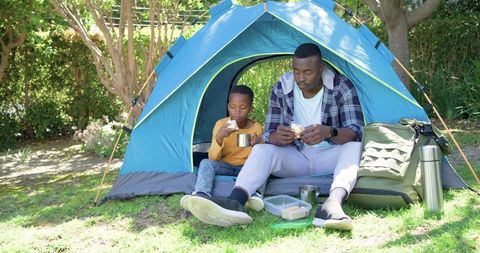 African american father and son camping at tent eating sandwiches sharing outdoor meal