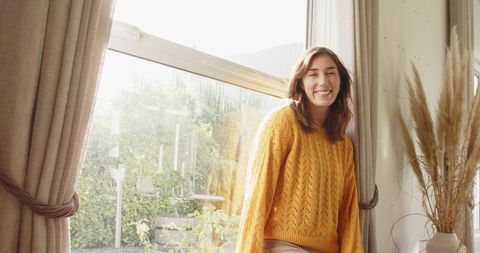 Sunlit mid adult woman smiling by sash window wearing mustard knit sweater, cozy home interior