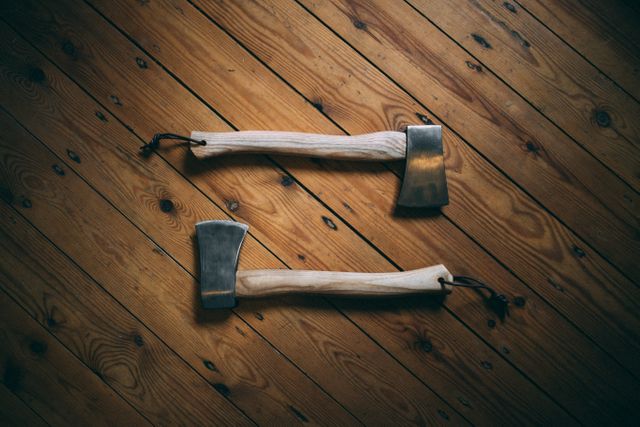 Two handcrafted axes resting parallel on a wooden floor featuring rich wood grain. Perfect for use in outdoor adventure blogs, woodworking magazines, craftsmen advertisements, or rustic decor themes.
