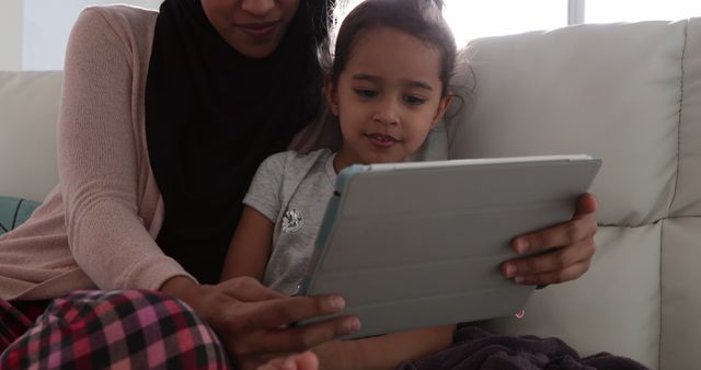 Front view of a young biracial woman wearing hijab with her young daughter in the sitting room, sitting on a sofa and using a tablet computer