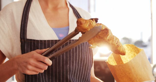 Baker wearing apron placing fresh croissant in paper bag with tongs. Ideal for illustrating bakery services, fresh pastries, breakfast foods, and small business operations.