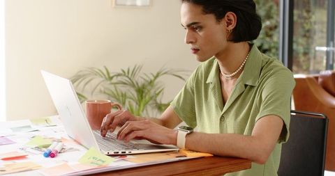 Young professional typing on laptop at home desk with sticky notes, coffee, and plants