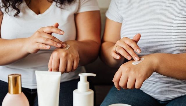 Two women applying hand cream on hands together on sofa for skincare self-care routine