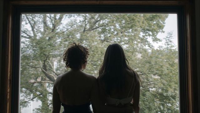 Backlit silhouettes of two women standing at large wooden window overlooking green trees