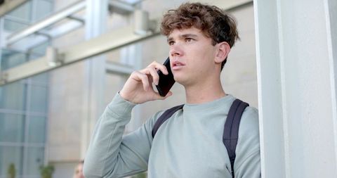 Young man calling on smartphone while standing at modern glass entrance with backpack
