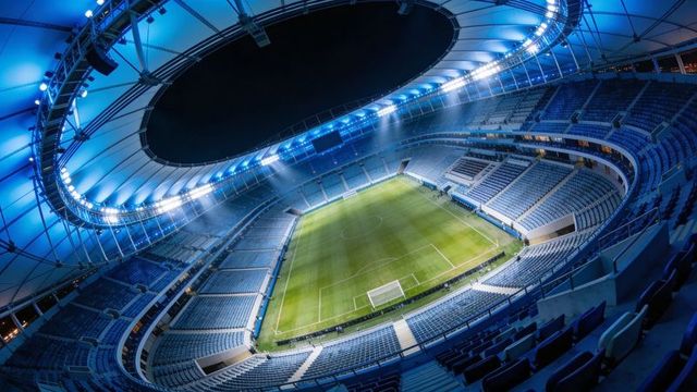 Blue-lit modern football stadium bowl illuminating pitch under oval roof at night