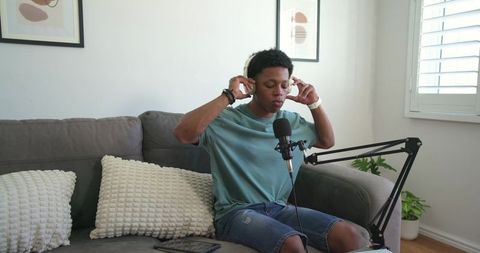 Podcaster adjusting headphones on sofa preparing to record with microphone and tablet in bright home
