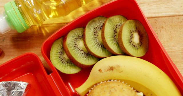 Juice bottle and slices of kiwi, banana and cupcake in red breakfast box on wooden counter. Food and nutrition, school time and childhood, vitamins and health care.