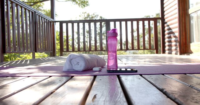 Yoga mat set up on a wooden deck in a natural outdoor setting, with a water bottle, towel, and phone placed on it. Useful for promoting outdoor yoga classes, fitness, and healthy lifestyle products. Ideal for blogs, websites, and advertisements focused on health and wellness.