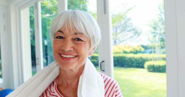 A senior Caucasian woman smiles warmly at the camera, with a towel around her neck suggesting she's engaged in physical activity or completed a workout, with copy space. Her cheerful expression and active lifestyle convey a positive message about health and fitness in later life.