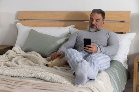 Mature man relaxing in bed petting puppy while scrolling smartphone in cozy bedroom