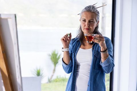 Senior artist drinking tea in sunlit studio contemplating next brushstroke in denim shirt