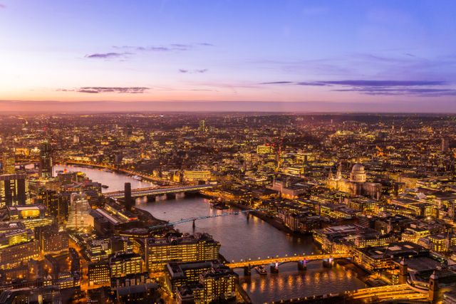 This image showcases an aerial view of London during sunset, with the River Thames meandering through the lit-up cityscape. Prominent landmarks like St Paul's Cathedral can be seen, along with several bridges crossing the river. This image can be used for travel promotions, city guides, educational materials about London, and articles related to urban development and infrastructure.