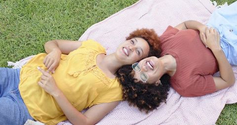 Mid-adult friends relaxing on pink picnic blanket laughing together on sunny green lawn