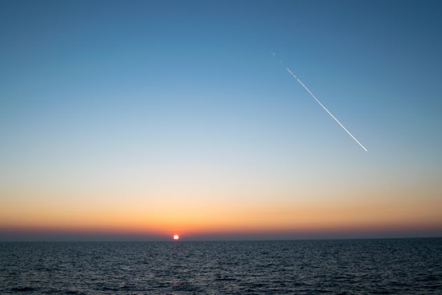 Peaceful sunset over ocean horizon with visible airplane contrail streaking across sky. Ideal for travel blogs, relaxation or meditation content, and serene scenery backgrounds.