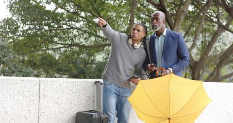 Traveler pointing while companion holding yellow umbrella and luggage at urban plaza