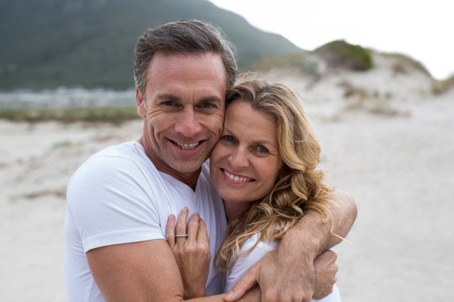 Mature couple smiling and embracing each other while standing on a beach with mountain in background. They are both wearing white t-shirts and appear happy and in love. This image could be used for promoting romantic getaways, lifestyle blogs, or advertisements emphasizing love and togetherness.