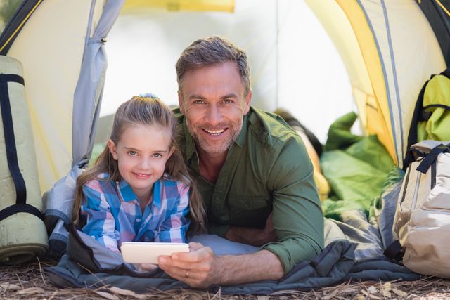 Father and daughter enjoying quality time together in a tent at a campsite. They are using a mobile phone, indicating a blend of nature and technology. Ideal for promoting family bonding, outdoor activities, camping gear, and technology use in nature.