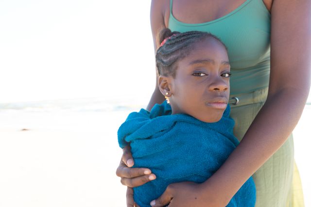 Midsection of african american mother wrapping daughter with towel while standing at beach. Copy space, unaltered, family, together, vacation, childhood, portrait, enjoyment and nature concept.
