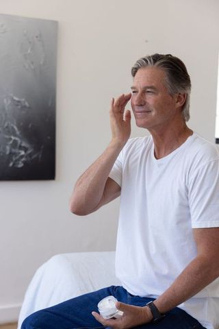 Senior man applying facial cream while sitting on bed wearing white t-shirt and wristwatch