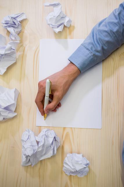 Business executive writing on blank paper surrounded by crumpled sheets on a wooden desk. Ideal for concepts related to brainstorming, creativity, problem solving, office work, and planning. Useful for business presentations, articles on productivity, and creative process illustrations.