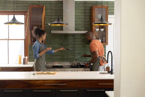 Couple cooking in modern kitchen with green tile backsplash, stainless-steel stove, pendant lights
