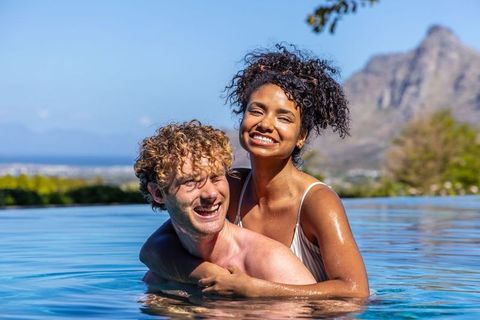 Smiling mixed-race couple enjoying infinity pool piggyback with mountain and ocean view