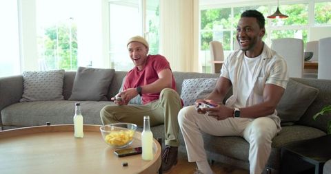 Playing Console Games on Couch Laughing Friends with Gamepads and Snacks in Sunlit Living Room