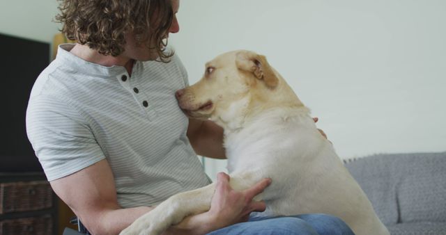A man is cuddling with his Labrador Retriever while sitting on a sofa at home. This scene shows the strong bond and affection between a pet and its owner. Useful for depicting pet ownership, companionship, and domesticity. Ideal for pet care blogs, advertisements for pet products, and lifestyle content.