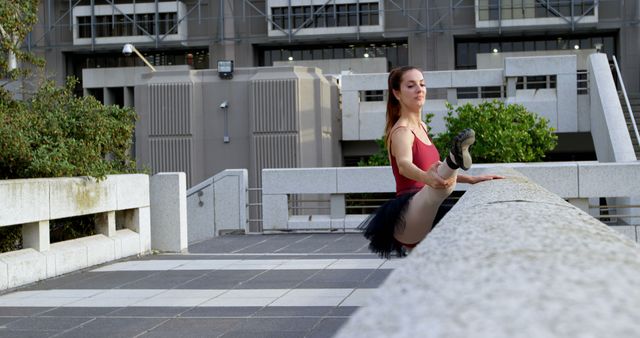 Ballet dancer stretching on a concrete railing in an urban setting, dressed in leotard and tutu. Useful for content related to dance, fitness, performing arts, urban exercises, and dedication in artistic practices.