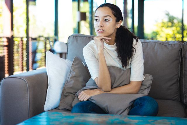 Thoughtful biracial young woman with hand on chin looking away while sitting on sofa at home - Download Free Stock Photos Pikwizard.com