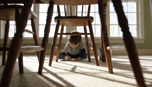 Young boy drawing under wooden dining table with crayons in sunlit room