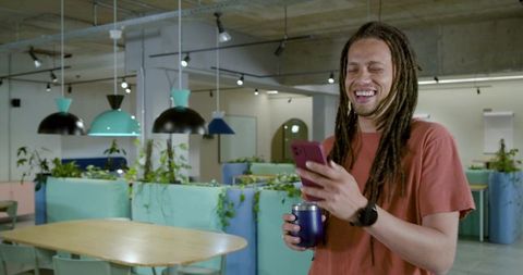 Laughing african american man using smartphone and tumbler in modern coworking lounge