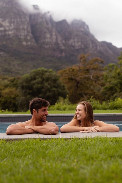 Couple enjoying a relaxing moment in a swimming pool with a scenic mountain backdrop. Ideal for use in travel brochures, vacation advertisements, lifestyle blogs, and romantic getaway promotions. Highlights themes of relaxation, nature, and togetherness.
